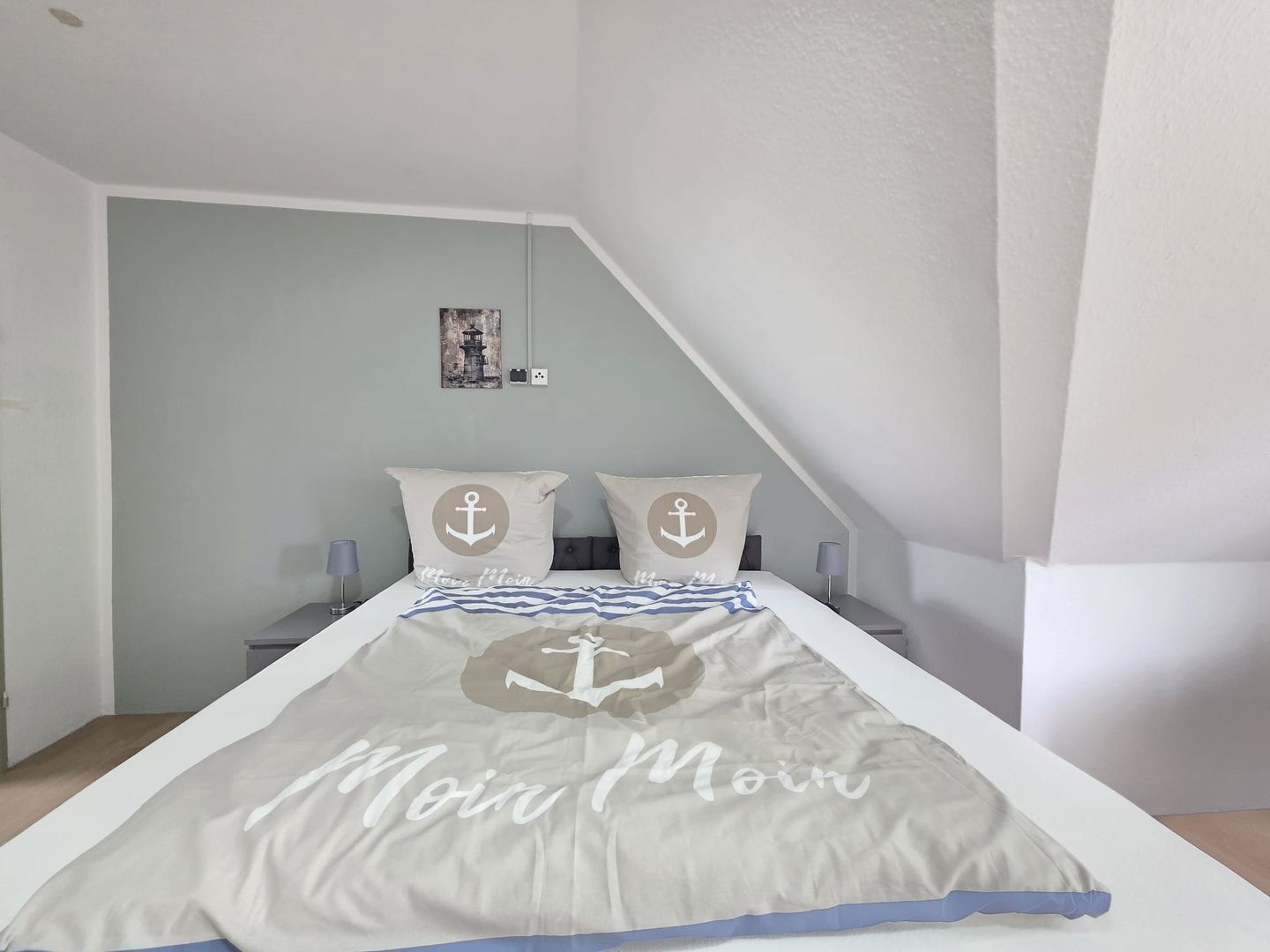 Ein Schlafzimmer mit einem Bett, zwei Kissen mit Anker-Motiven, einem Nachttisch mit Lampe und einem Wandbild.