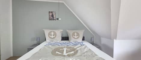 Un dormitorio con una cama, dos almohadas con motivos de anclas, una mesita de noche con una lámpara y un cuadro colgado en la pared.