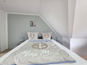 Een slaapkamer met een bed, twee kussens met ankerprint, een nachtkastje met een lamp en een aan de muur hangend schilderij.