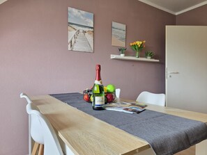 A dining area with a wooden table, white chairs, a bottle of champagne, a bowl of fruit, and two framed pictures on the wall. - Vacation apartment Kliffkannte - Holiday apartment Kliffkannte - Modern time-out at the (Norden)