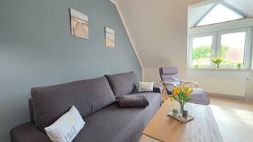 Ein modernes Wohnzimmer mit einem grauen Sofa, einem Holztisch und Blick auf Grün hinter einem großen Fenster.