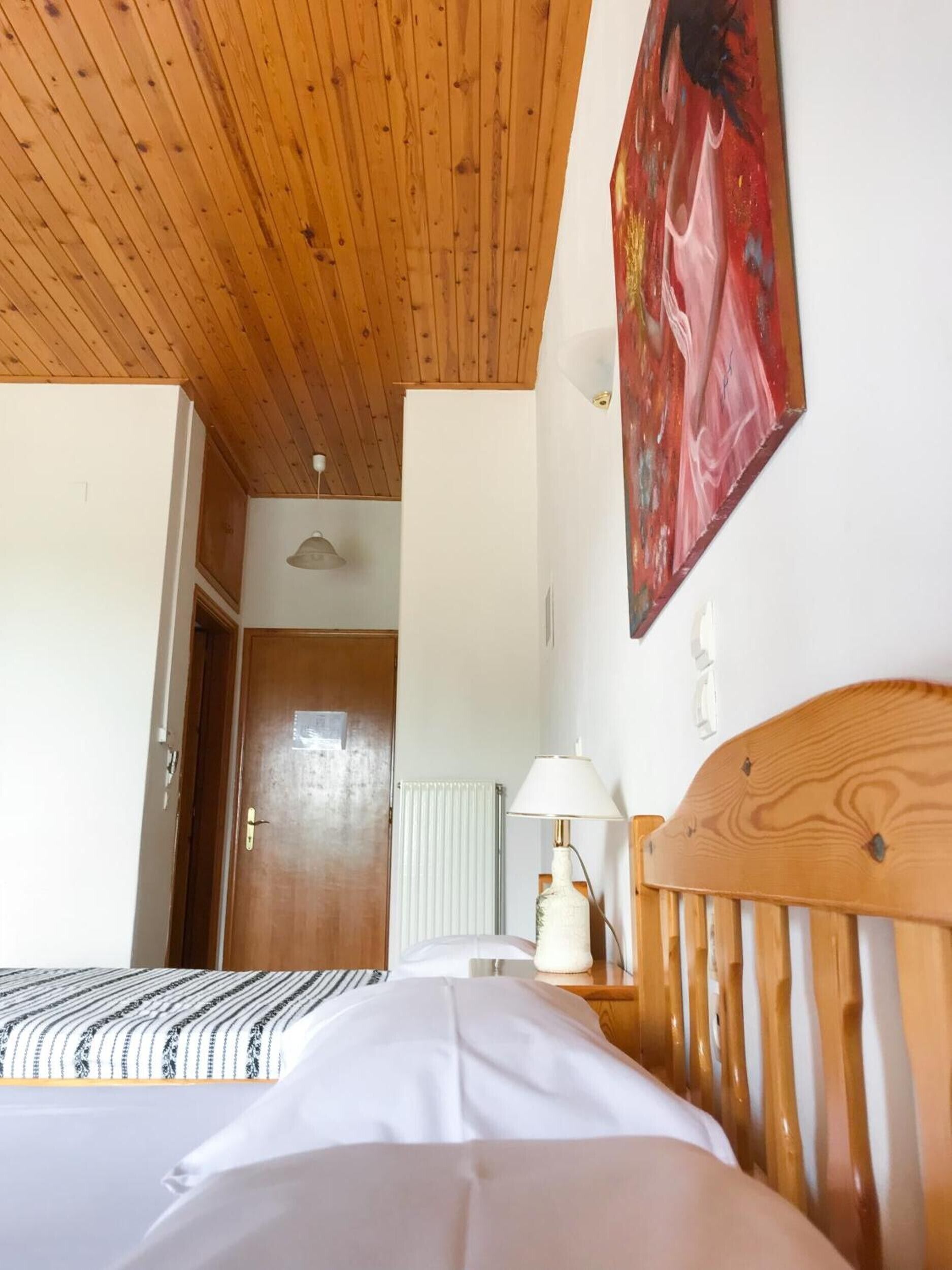 A bedroom with a wooden ceiling, a bed with white bedding, a nightstand with a lamp, and a door leading to another room.