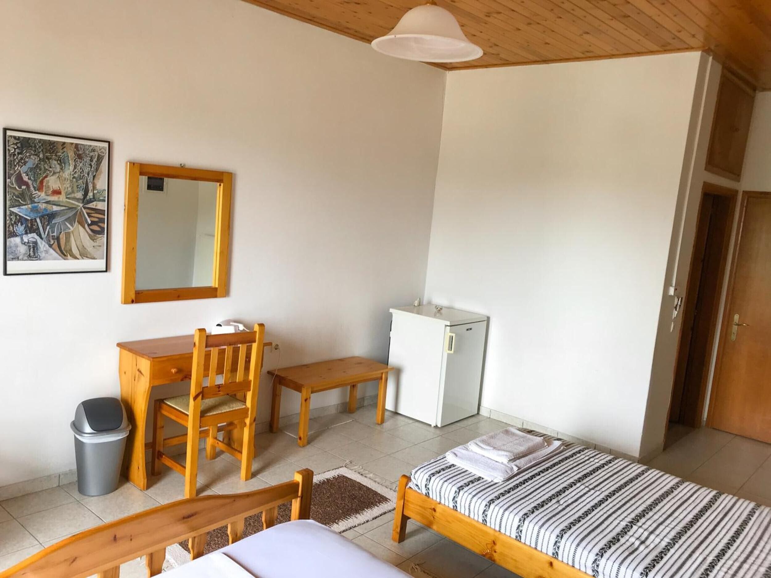 A bedroom with a bed, a desk, a chair, a small refrigerator, a trash bin, and a mirror.