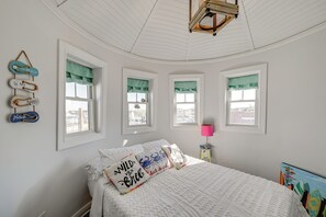 Quarto com uma cama branca, três janelas com cortinas verdes, um ventilador de teto e decorações nas paredes incluindo uma placa e um abajur.