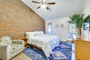 Une chambre avec un mur en briques, un lit avec une literie blanche, un tapis à motifs bleus, un ventilateur de plafond et une plante en pot.