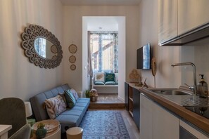 Ein kompakter Wohnbereich mit Sofa, einer kleinen Küchenzeile, einem Esstisch und Blick durch ein Fenster.