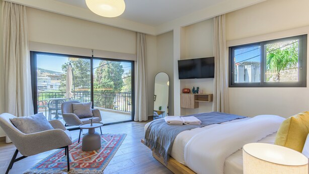Un dormitorio con una cama grande, televisión, balcón con vistas y zona de estar con sillas.