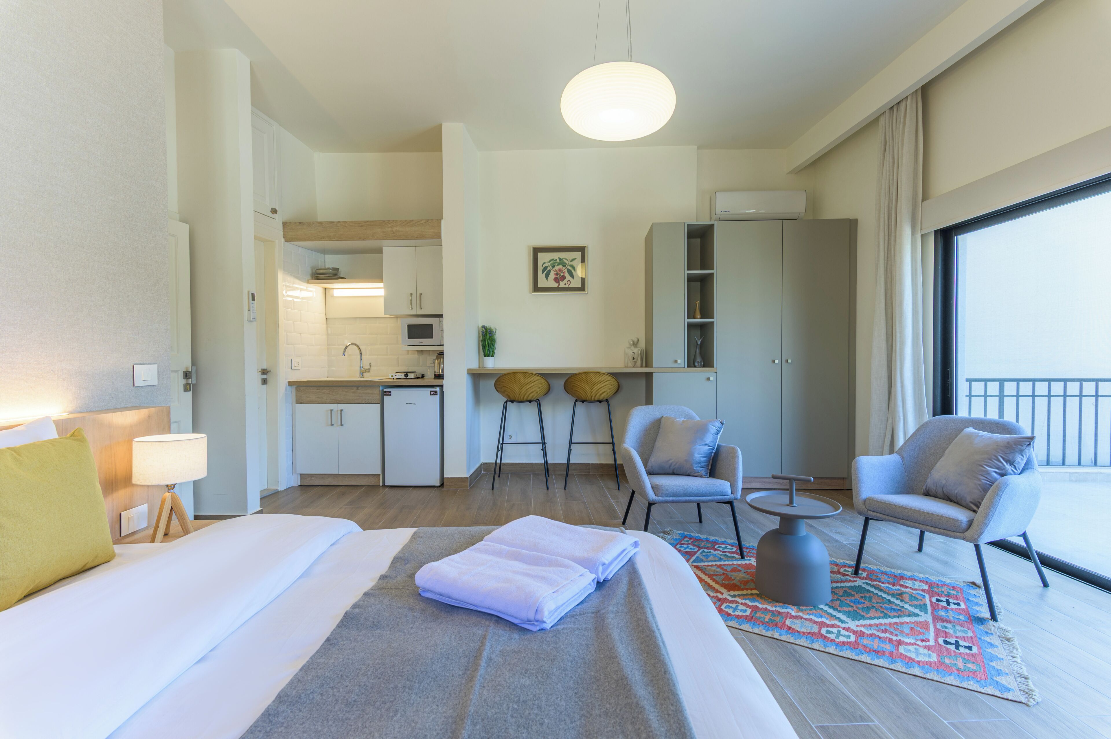 Hotellihuone, jossa on sänky, työpöytä, tuoli, pieni keittotila ja suuri ikkuna.