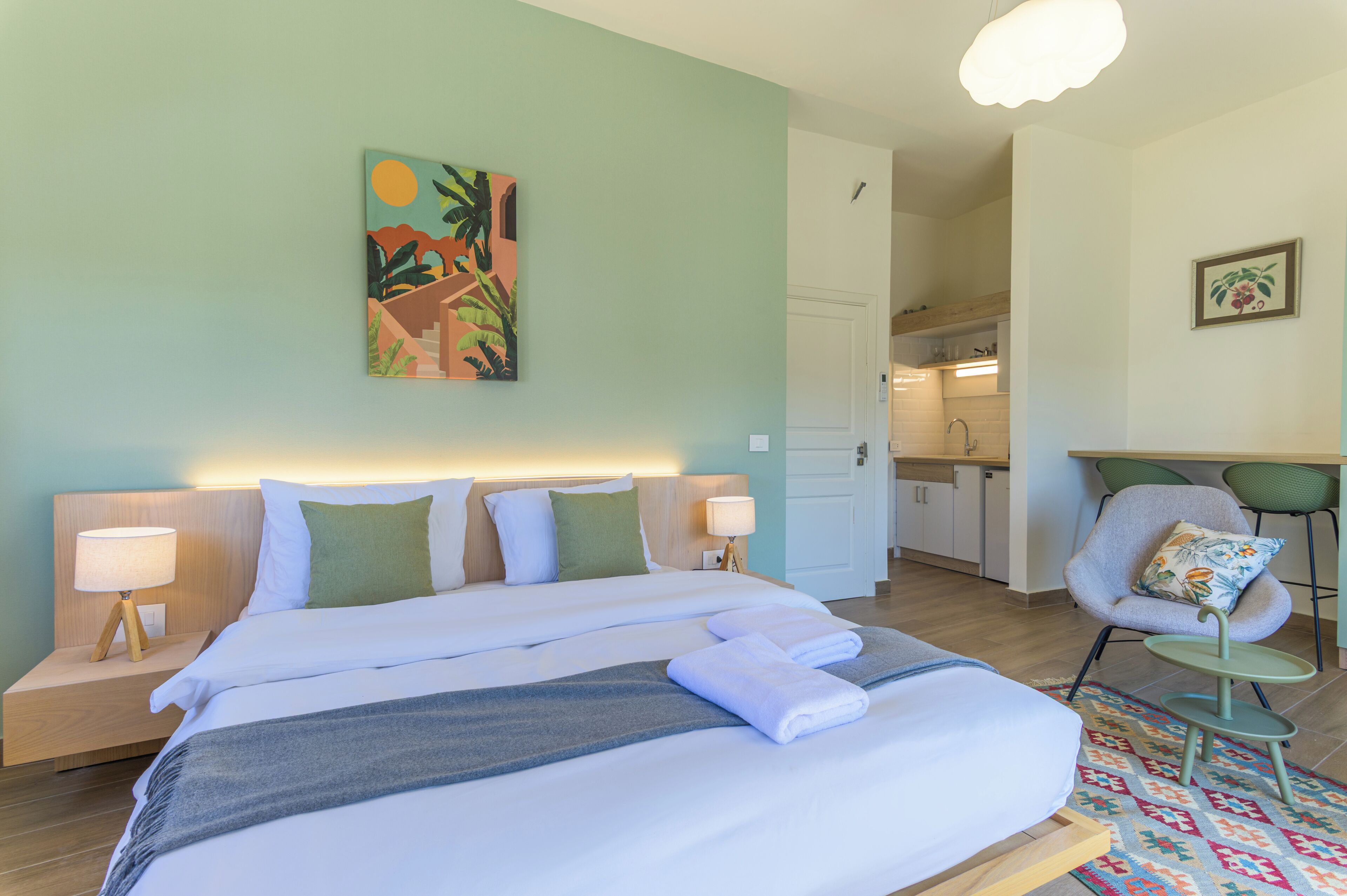 A hotel room with a bed, bedside tables, a chair, a desk, and a kitchen area.