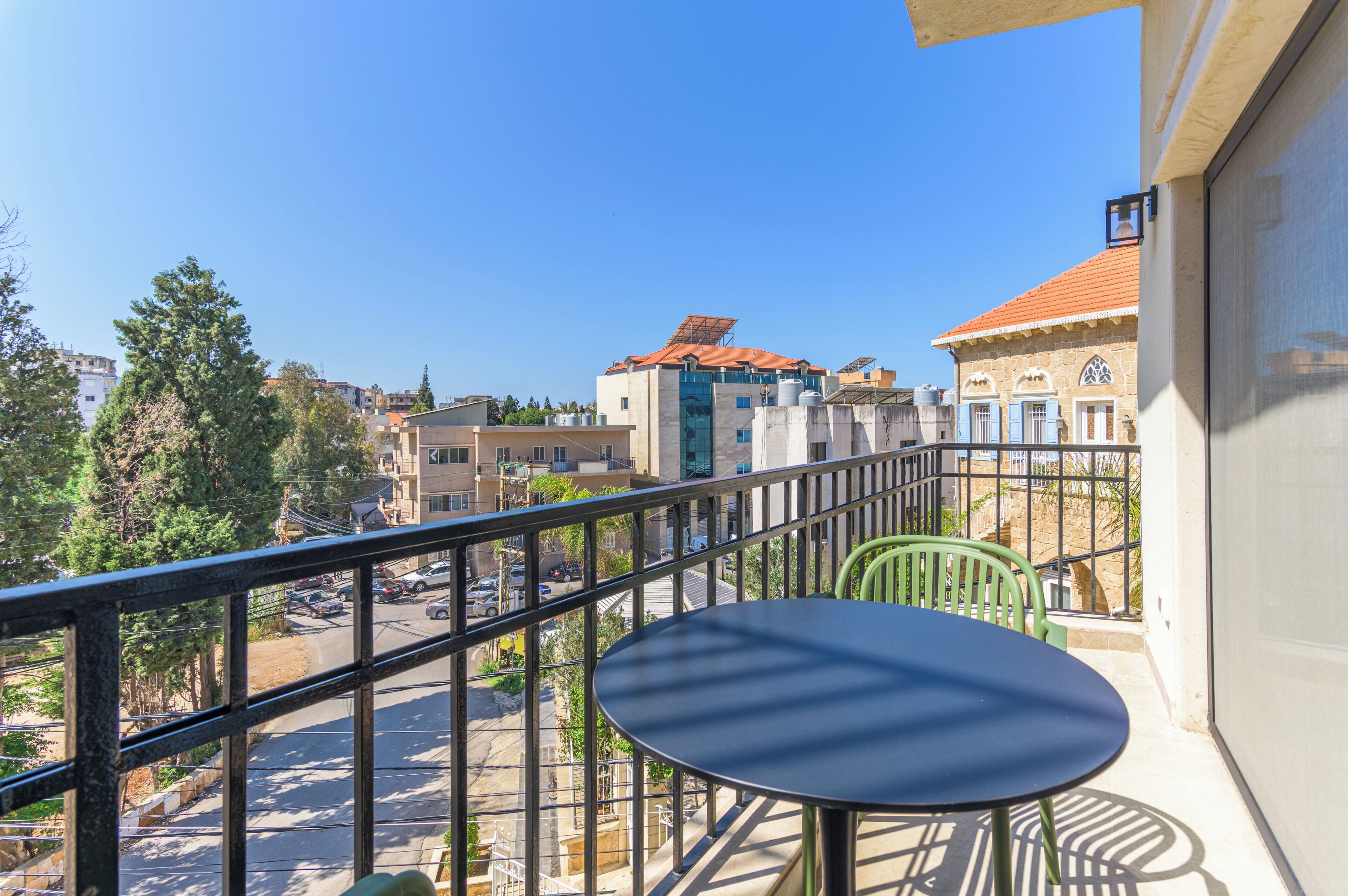 A balcony with a table and chairs, overlooking a street with buildings and trees.