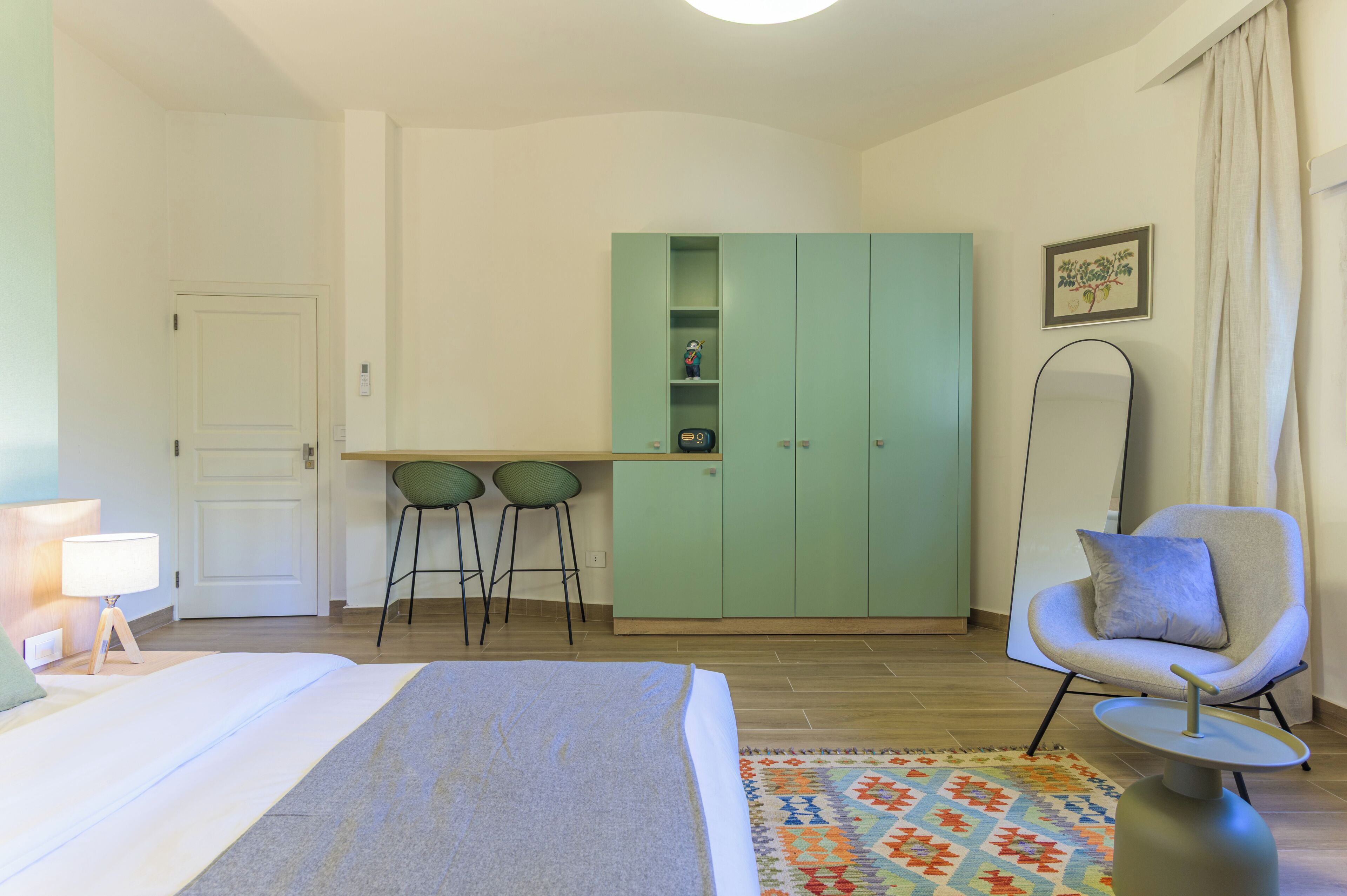 A bedroom with a bed, a desk, two chairs, a wardrobe, a chair, a lamp, and a rug.