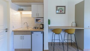 Una cocina compacta con armarios blancos, un microondas, un lavamanos y un refrigerador pequeño. Hay dos sillas amarillas y una planta en maceta sobre la encimera.