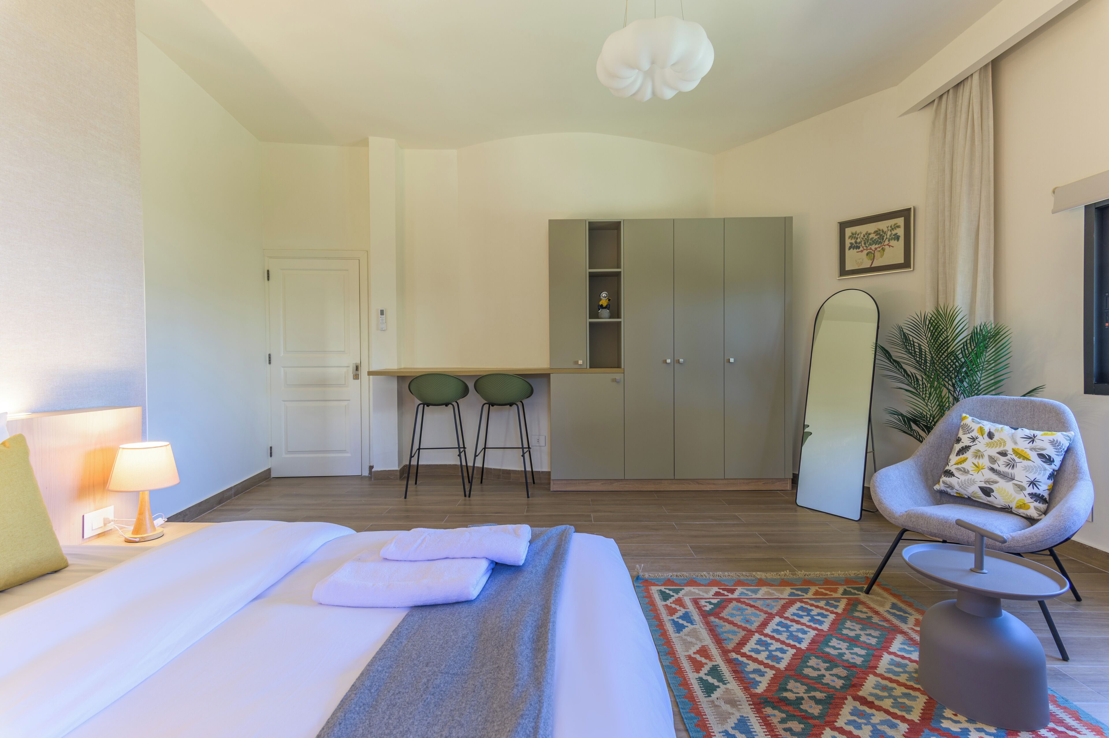 Ein Hotelzimmer mit einem Bett, einem Schreibtisch mit zwei Stühlen, einem Kleiderschrank, einem Sessel mit Kissen und einer Pendelleuchte.