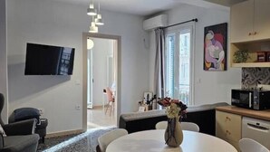 A modern living room with a flat-screen TV, a dining table with chairs, a kitchen area with a microwave, and a large window with curtains. - Zante Cozy Apartment (Zakinthos)