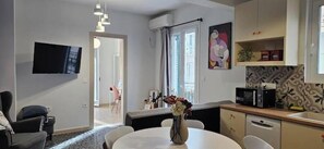 Ein modernes Wohnzimmer mit Flachbildfernseher, einem Esstisch mit Stühlen, einer Küchenzeile inklusive Mikrowelle und einem großen Fenster mit Vorhängen.