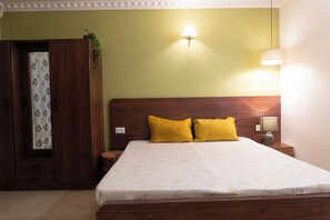 A bedroom with a wooden bed, two yellow pillows, a nightstand with a lamp, and a wardrobe with a floral curtain. - HomeSlice Koramangala (Bengaluru)