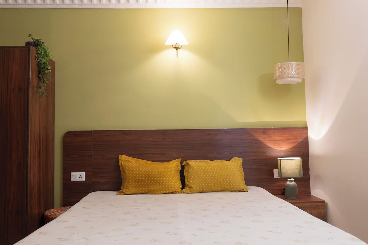Een slaapkamer met een houten hoofdbord, twee gele kussens, een bedlamp en een wandlamp.