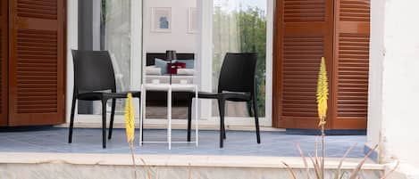 Un área de estar moderna al aire libre con dos sillas y una mesa, una escalera que sube a un nivel superior y vistas de un edificio con un balcón.