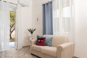 A room with a beige sofa, a small table with a plant, a blue curtain, and a window with a view of trees. - Villa Comar (Forio)