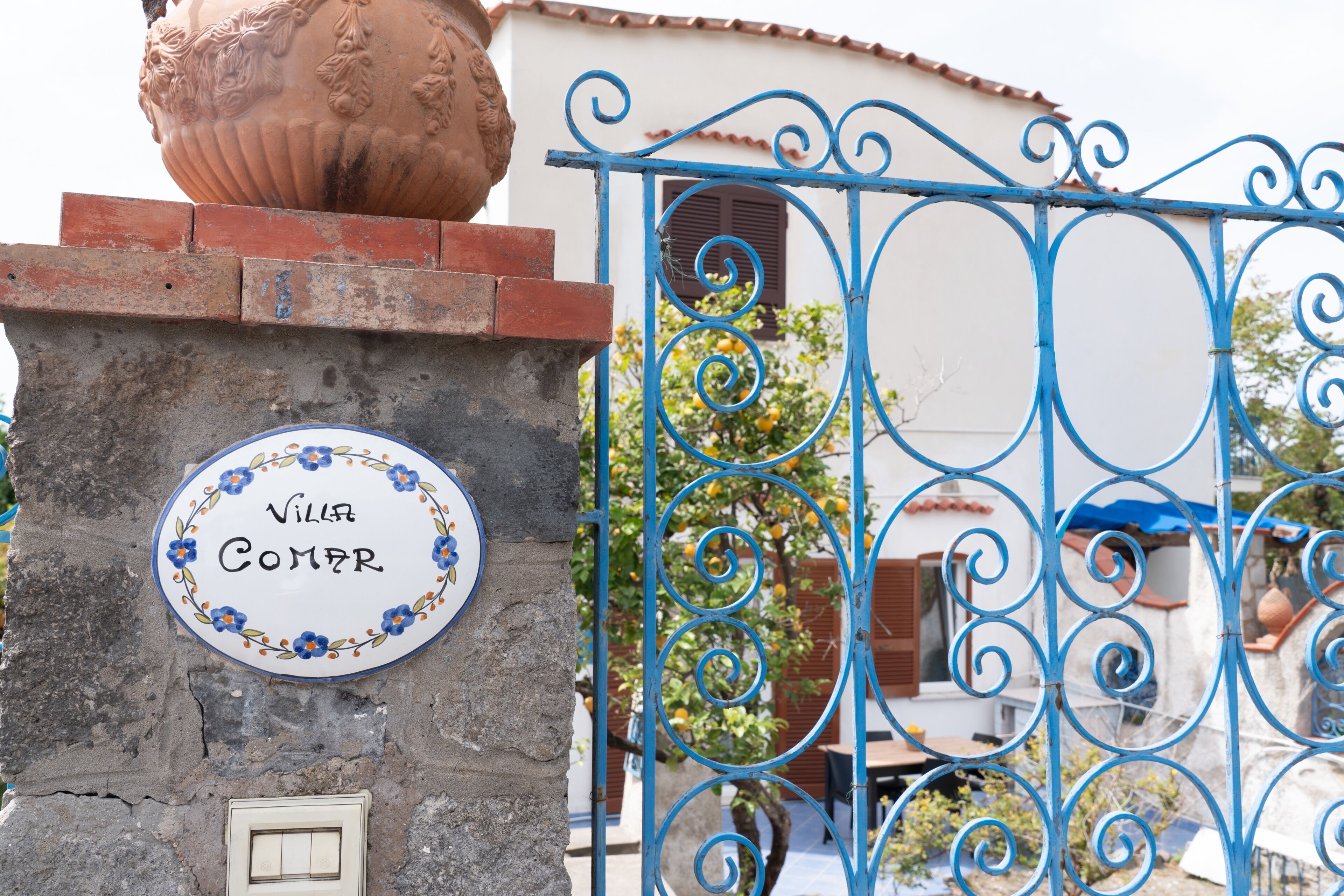 Una villa llamada 'Villa Comer' con un letrero decorativo, una maceta de barro grande y una verja de hierro azulado.