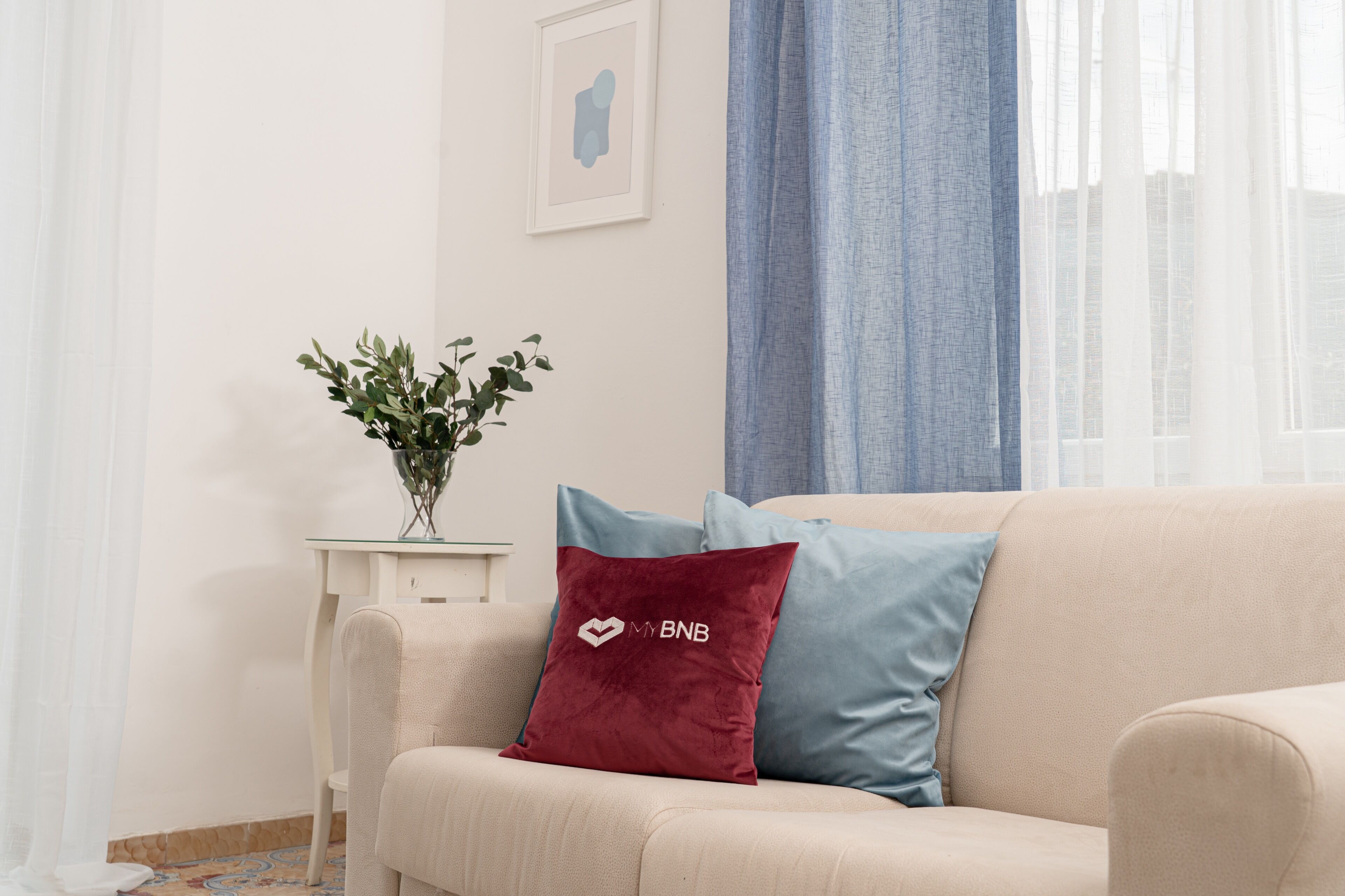 Een woonkamer met een beige bank, een rode kussen met een logo, een blauw kussen, een kleine tafel met een plant en een ingelijste foto aan de muur.