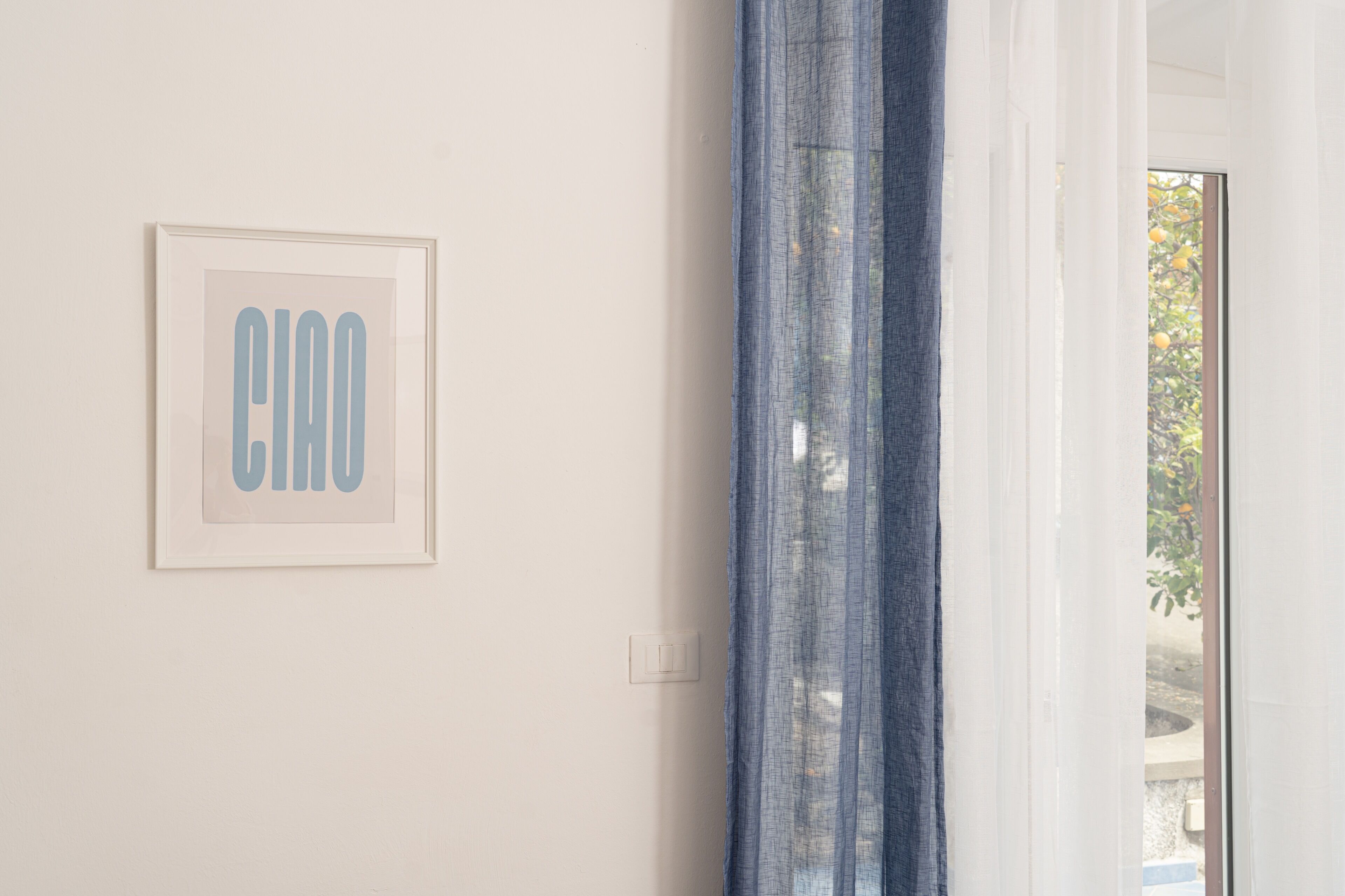 Une œuvre d’art encadrée avec le mot « Ciao » sur un mur uni, un rideau bleu partiellement tiré, et un aperçu d’un espace extérieur avec des arbres.