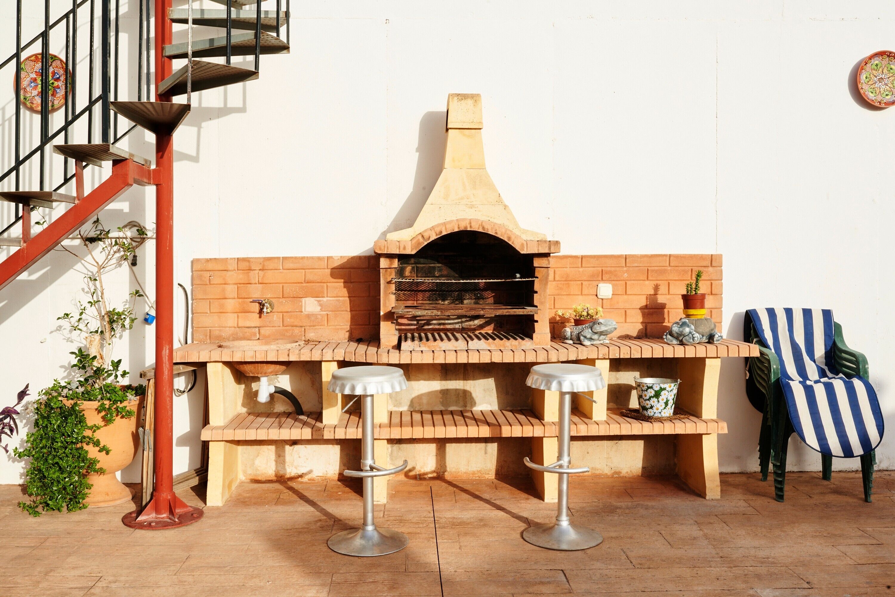 A brick outdoor kitchen with a chimney, two bar stools, a potted plant, and a chair.