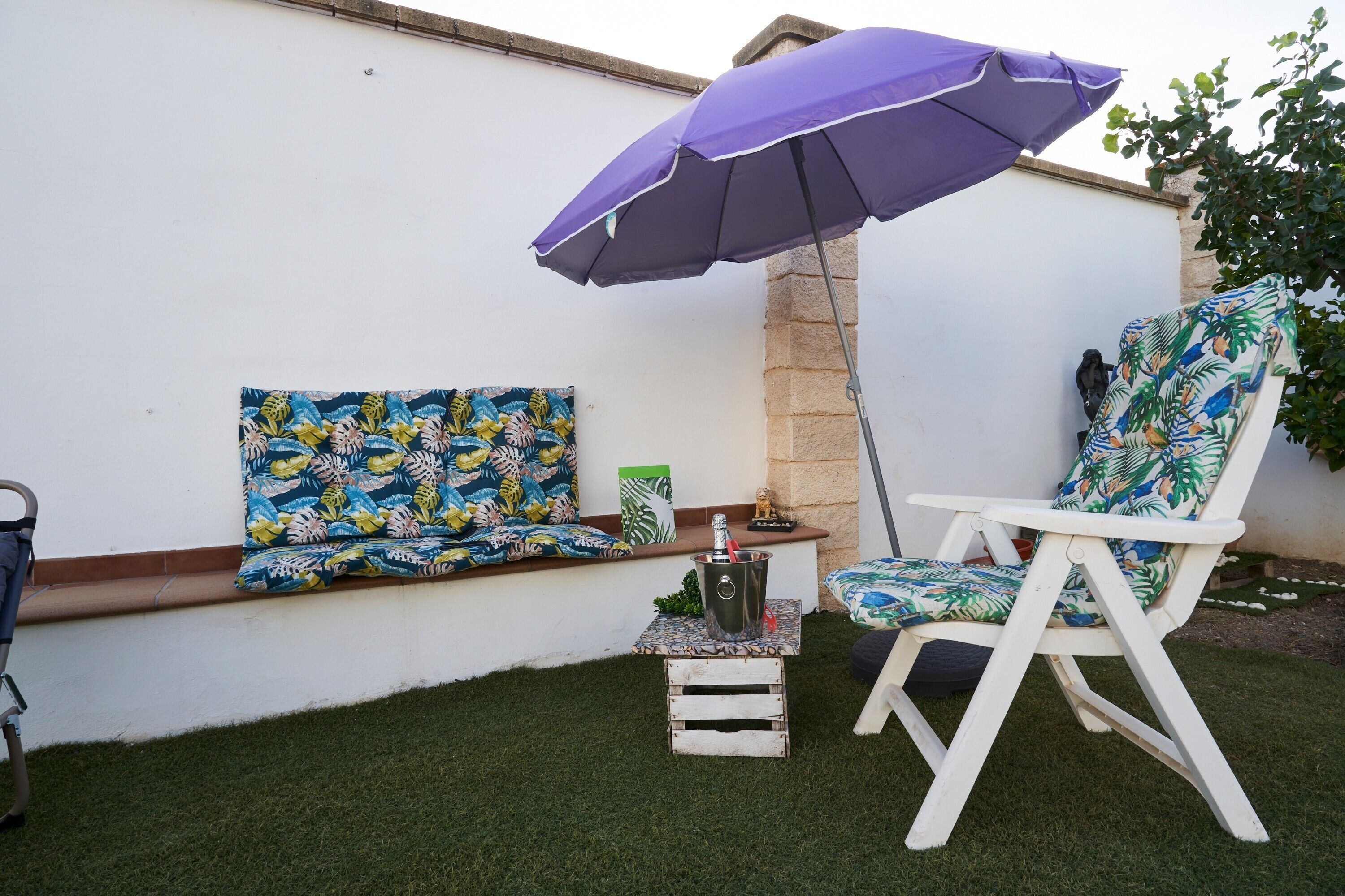 Un patio con una silla blanca, un banco con cojín floral, un paraguas púrpura y una pequeña mesa con una bebida.