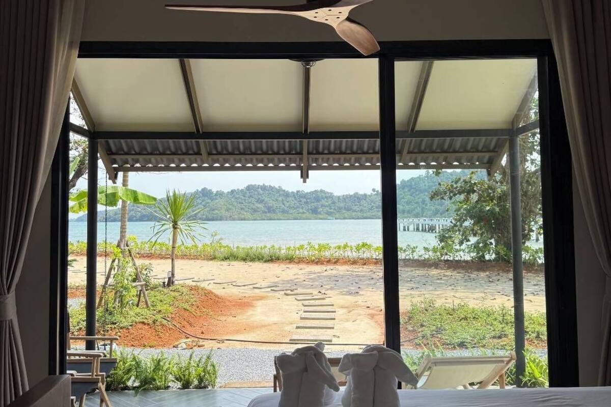 A bedroom with a bed, a ceiling fan, and a view of a beach and water.