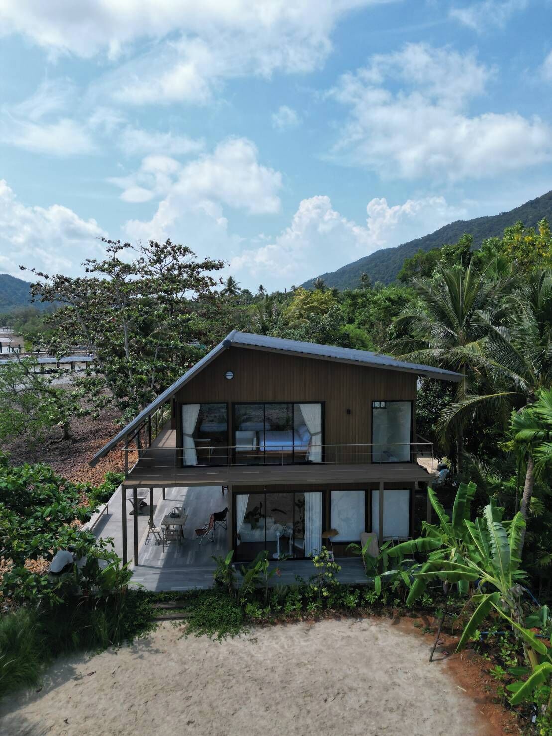 A modern two-story house with a balcony, surrounded by lush greenery and a mountainous backdrop.
