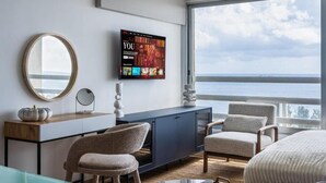 Habitación de hotel moderna con televisor grande, espejo redondo, consola de madera, dos sillas, cama y vista al mar.