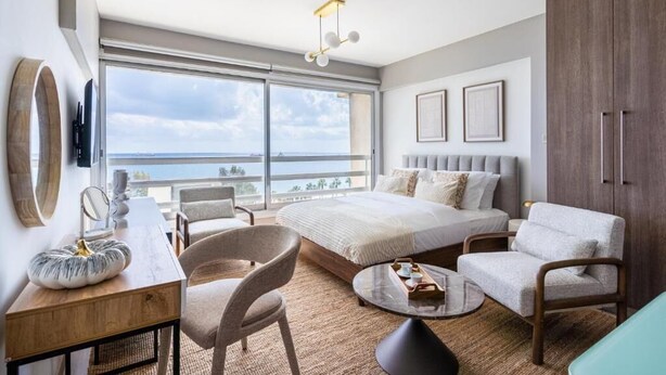 Una habitación de hotel moderna con una cama grande, un escritorio con una silla, una mesa de centro redonda y vista al mar.