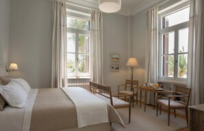 Ein Schlafzimmer mit einem Bett, zwei Stühlen, einem kleinen Tisch und zwei Fenstern mit Vorhängen.