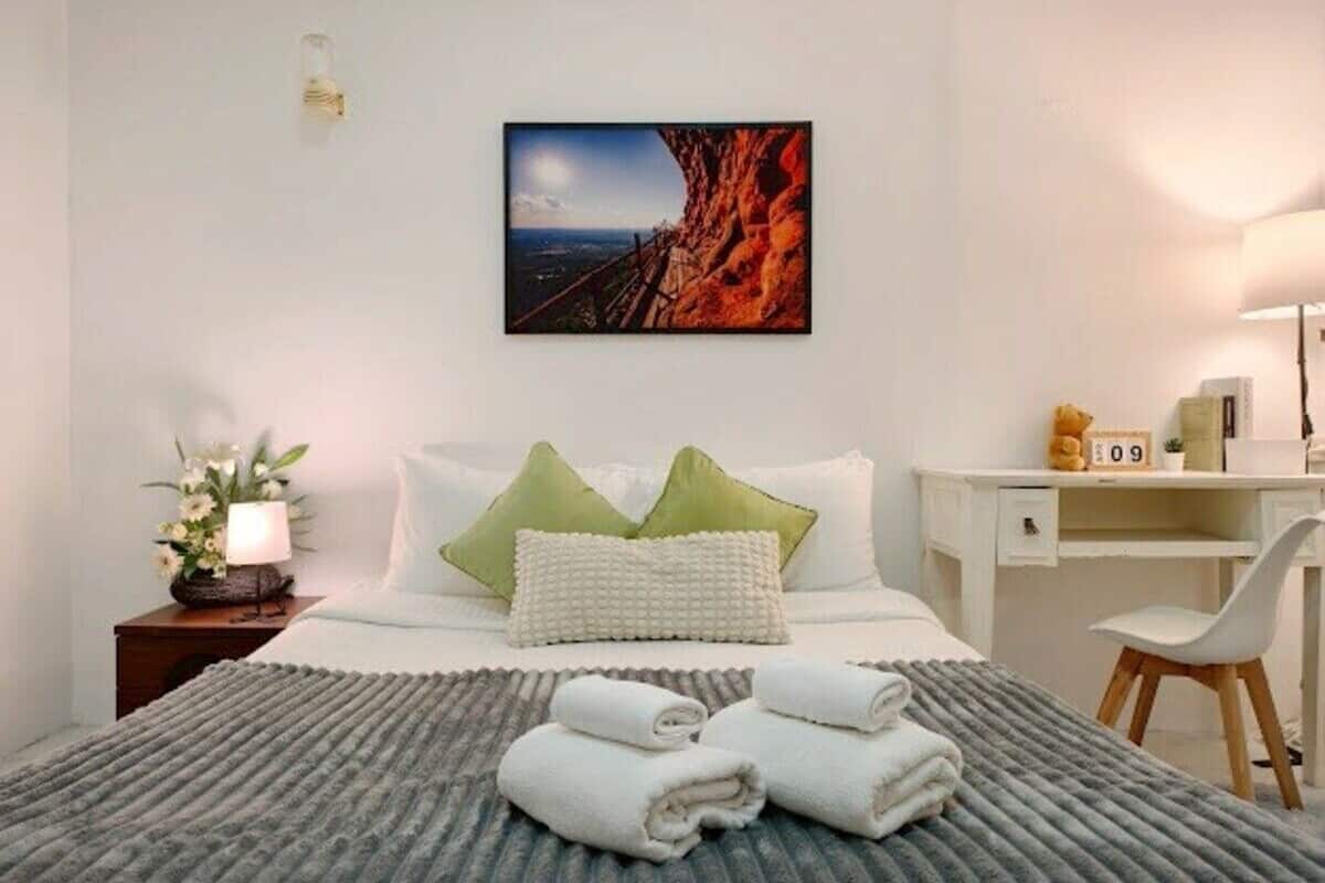 A hotel room with a bed, a desk, a chair, a nightstand, a lamp, a framed picture, and a clock.