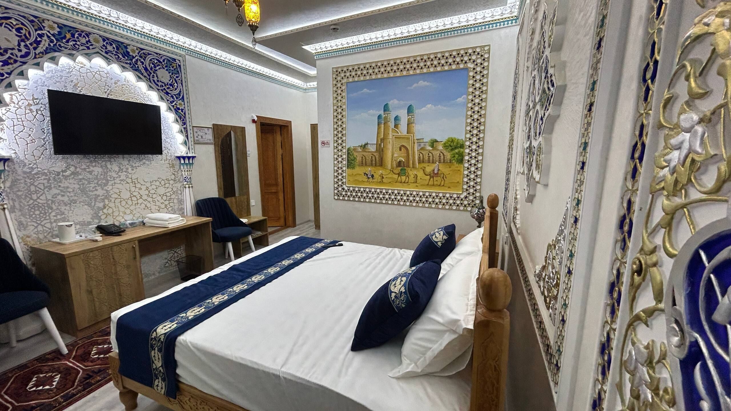 A hotel room with a bed, desk, chair, and a painting of a mosque on the wall.