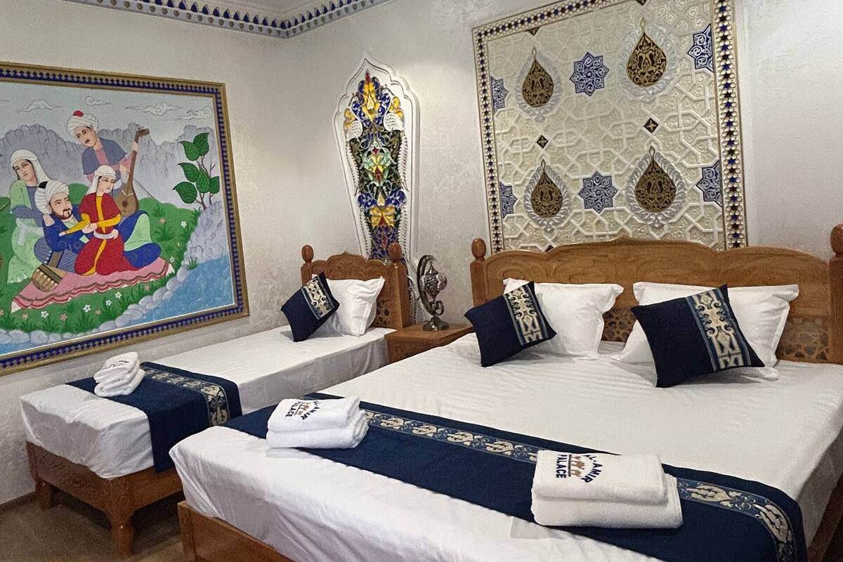 Quarto de hotel com duas camas de madeira, roupa de cama branca, almofadas decorativas, uma tapeçaria grande na parede e um tapete estampado.
