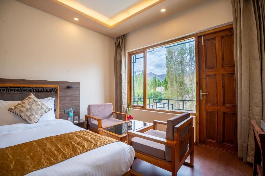 A hotel room with a bed, a small table with chairs, a window with a view of trees and mountains, and a wooden door.
