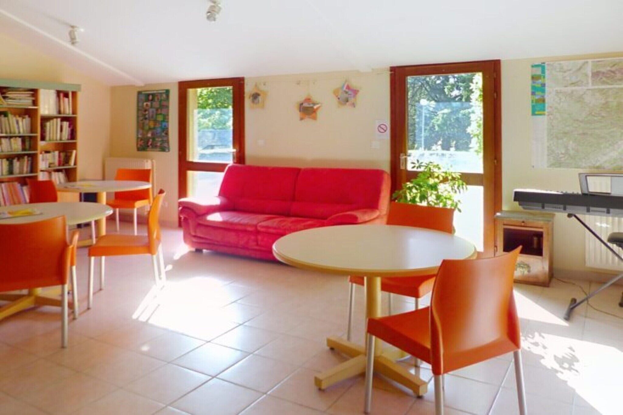 Ein Zimmer mit einem roten Sofa, orangenen Sesseln, einem runden Tisch, einem Bücherregal und einer Tastatur.