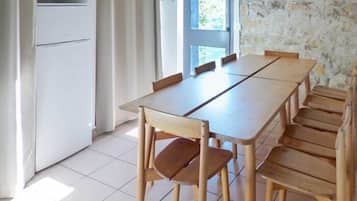 A dining area with a long wooden table and chairs, a white refrigerator, and a stone wall.