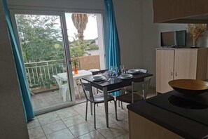 Comedor con una mesa lista para cuatro, zona de cocina con armarios y un televisor, y un ventanal corredizo que da a un espacio exterior con vistas a árboles y una valla.