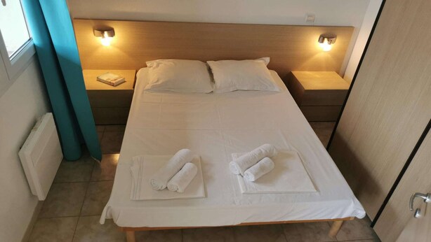 Een net opgemaakt bed met kussens, twee nachtlampjes en opgerolde handdoeken op het bed.