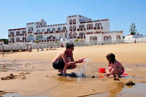 Een strand met een groot gebouw op de achtergrond, mensen die in het zand spelen en een heldere lucht.