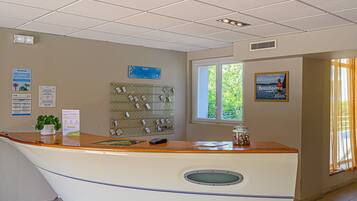 A reception desk with a wooden top, a plant, a bulletin board, and a window with a view of greenery.