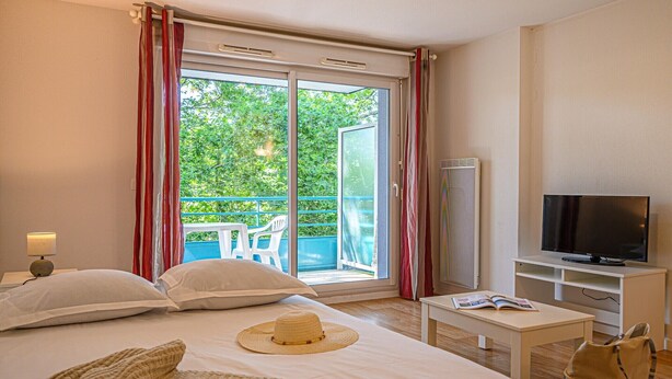 Habitación de hotel con cama, televisor, balcón con vistas a vegetación y ventana con cortinas.