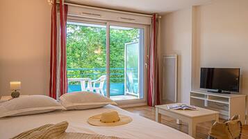 A hotel room with a bed, a TV, a balcony with a view of greenery, and a window with curtains.