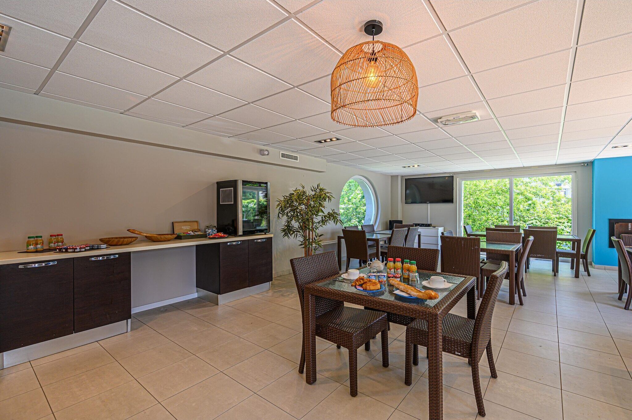 A modern kitchen with dark cabinetry, a dining area with wicker chairs, and a large window offering a view of greenery.