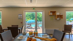 Un comedor con sillas de mimbre, una mesa de vidrio, alimentos para el desayuno y vistas a la piscina y la vegetación.