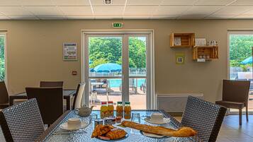 Un comedor con sillas de mimbre, una mesa de vidrio, alimentos para el desayuno y vistas a la piscina y la vegetación.