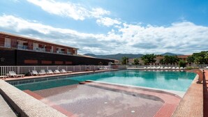 Piscina con sillas de descanso, zona cubierta y montañas al fondo.