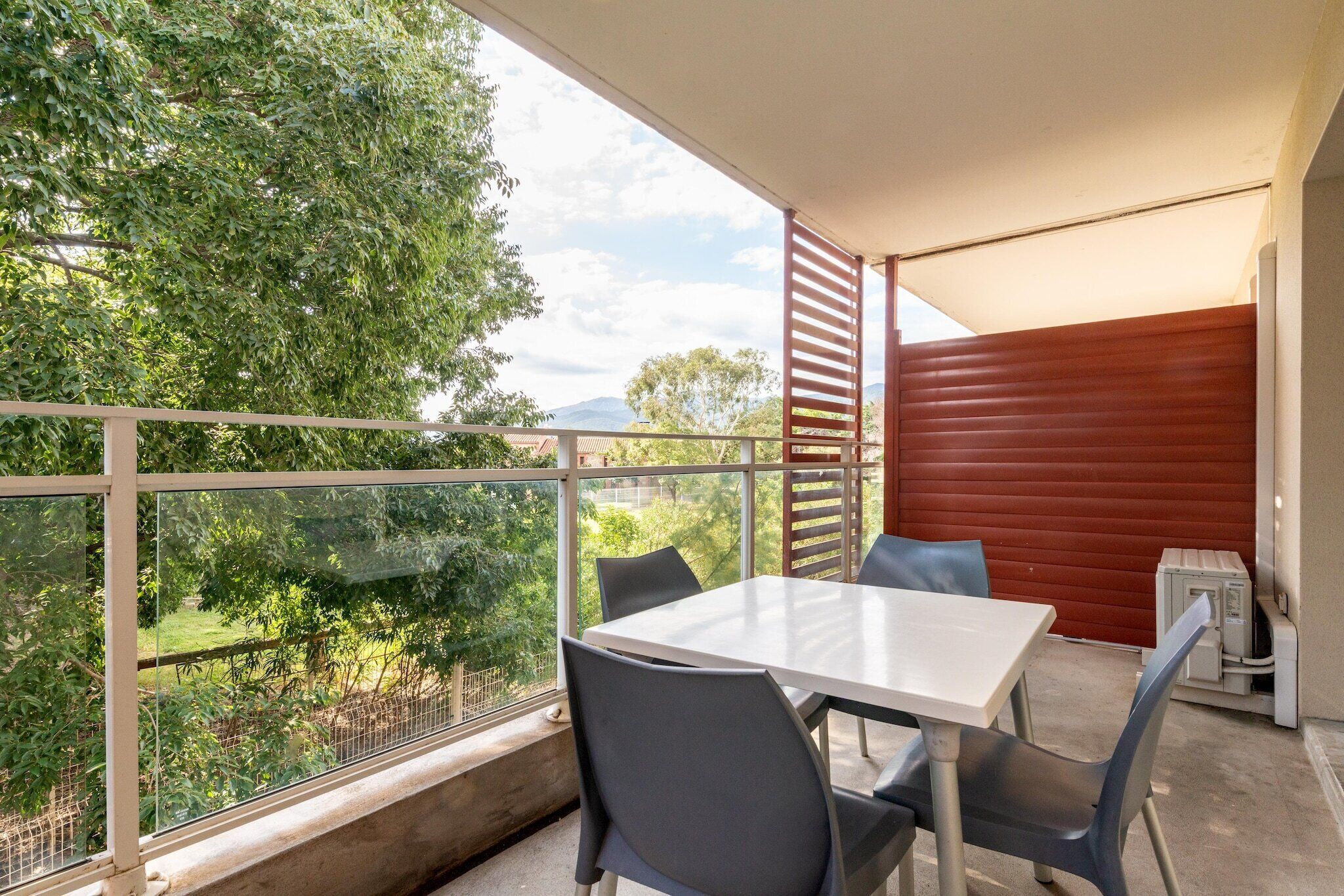 A balcony with a table and chairs, a red shutter, and a view of greenery and mountains.